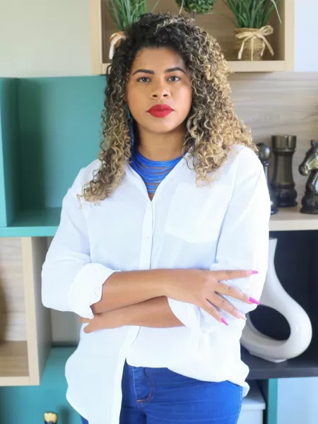 Daiane Mello é uma mulher negra de cabelos cacheados com pontas loiras. Ela veste uma camisa branca, colar azul e calça jeans. Daiane também usa um batom vermelho